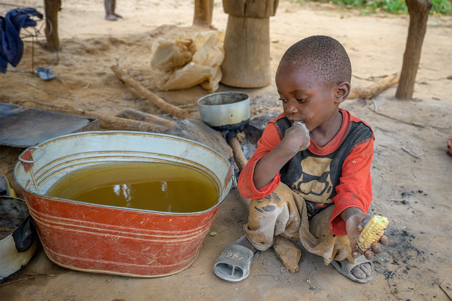 Wearing torn yellow pants and dusty sandals, a young boy squats in the dirt by a metal tub of dirty water in Zambia.