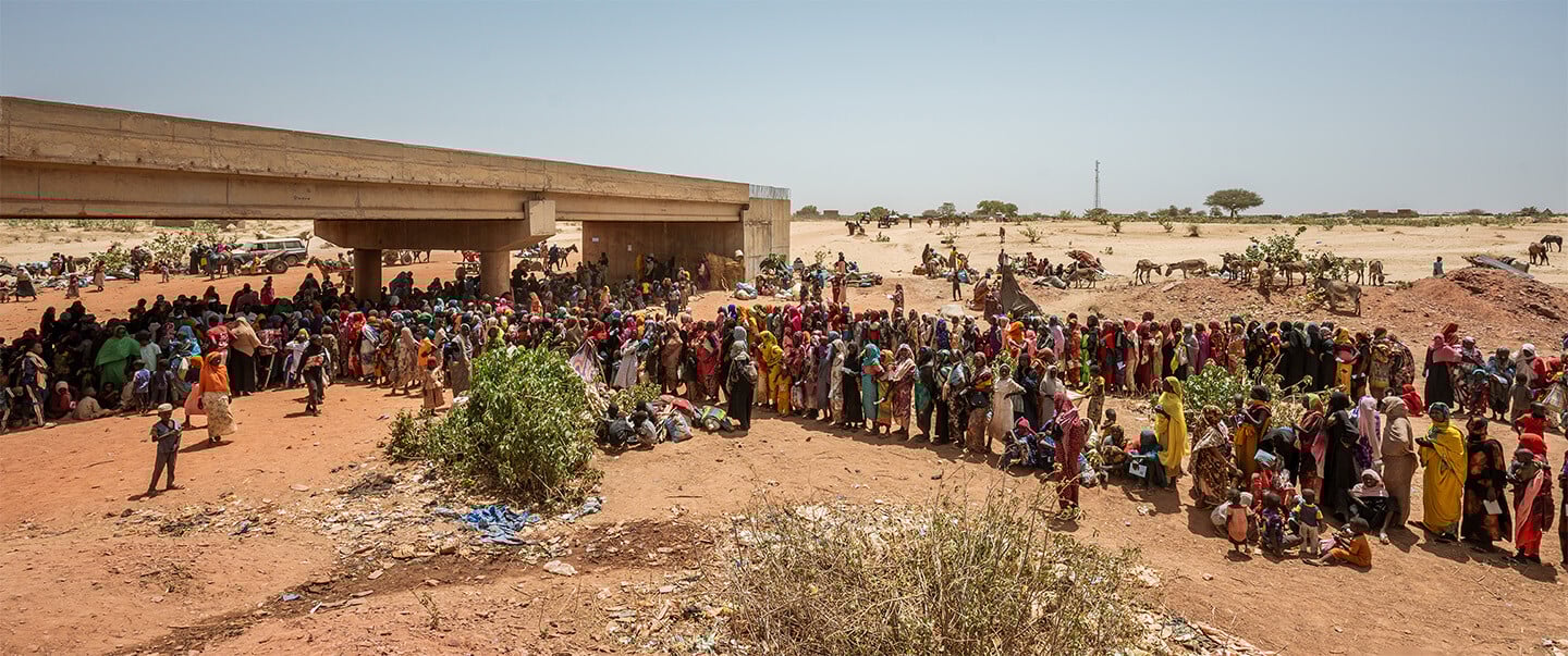 Under an incomplete bridge in the desert, a massive crowd of people, mostly women and children, wait in line to be allowed entry as refugees.