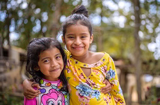 Two girls in Honduras smile with their arms around each other’s shoulders.
