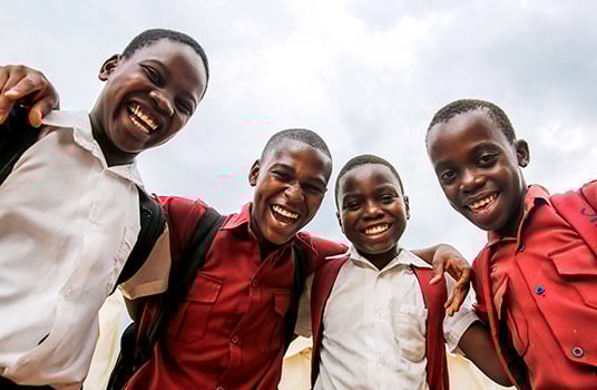 Four boys wearing red and white school uniforms smile widely with their arms around each other.