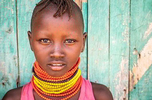 A young girl from Turkana, Kenya, wearing stacked red, yellow, and black beaded necklaces has a thoughtful expression.