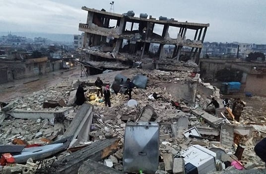 People walk on heaps of rubble in front of the shell of a building ruined from the earthquake in northwestern Syria.