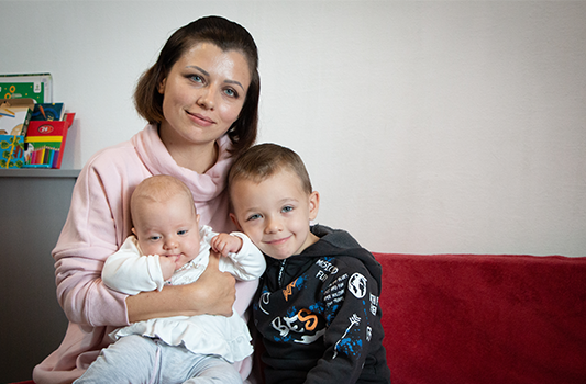 A woman wearing a pink sweater gently smiles with her 4-month-old daughter on her lap and 6-year-old son leaning against her.