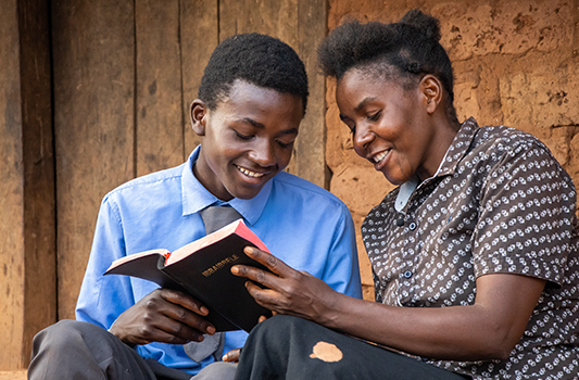 A teen boy and his mother smile while reading a Bible and sitting together outside a home in Zambia.
