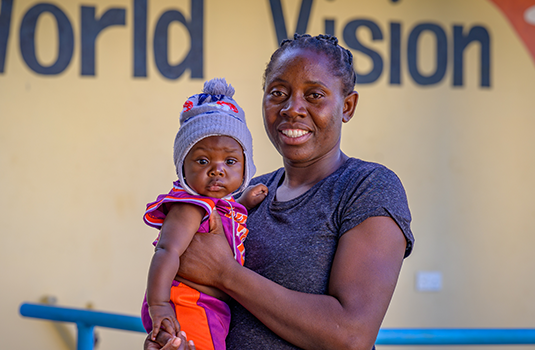 A smiling woman holds a baby in front of a wall that reads “World Vision”