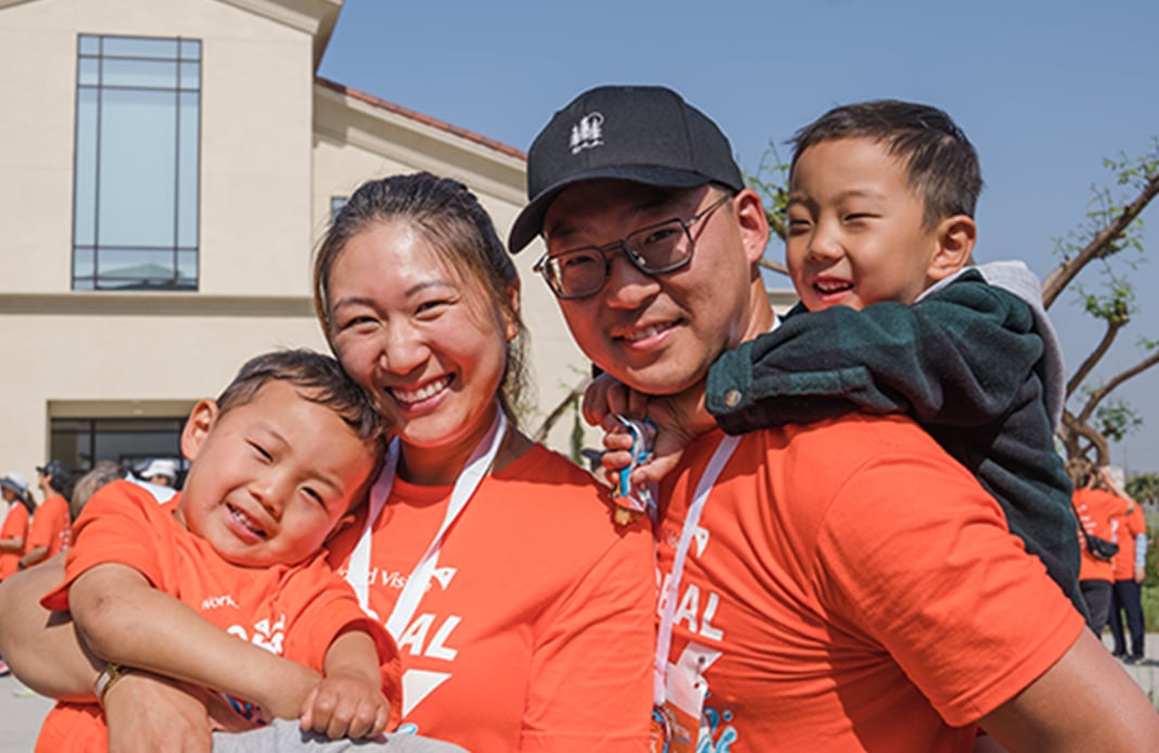 A woman, a man, and two young boys smile in front of a church. Three of them wear orange Global 6K for Water T-shirts.
