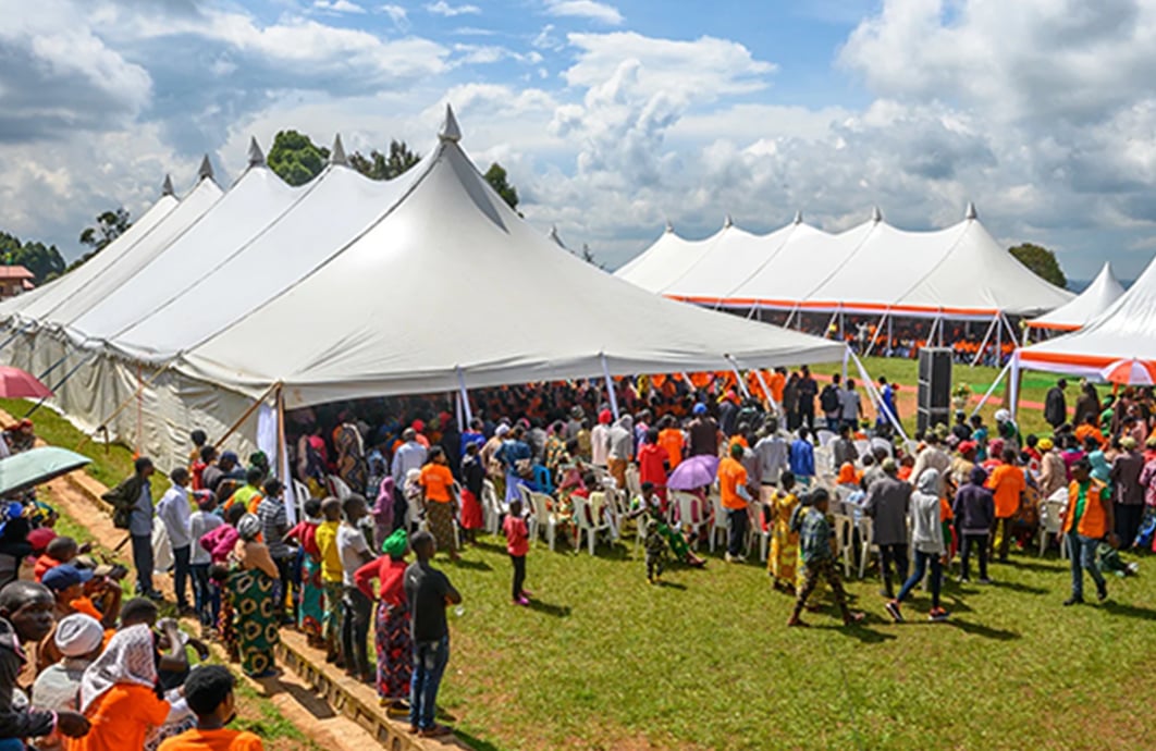 A large group of people in Rwanda stand next to and under large tents in a field.