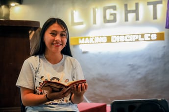A teenage girl in the Philippines sits in her church, holding her Bible and smiling.