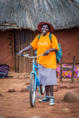A smiling community health worker comes to the local clinic in Zimbabwe, riding her blue bike, wearing an orange World Vision shirt and a large-brimmed hat .