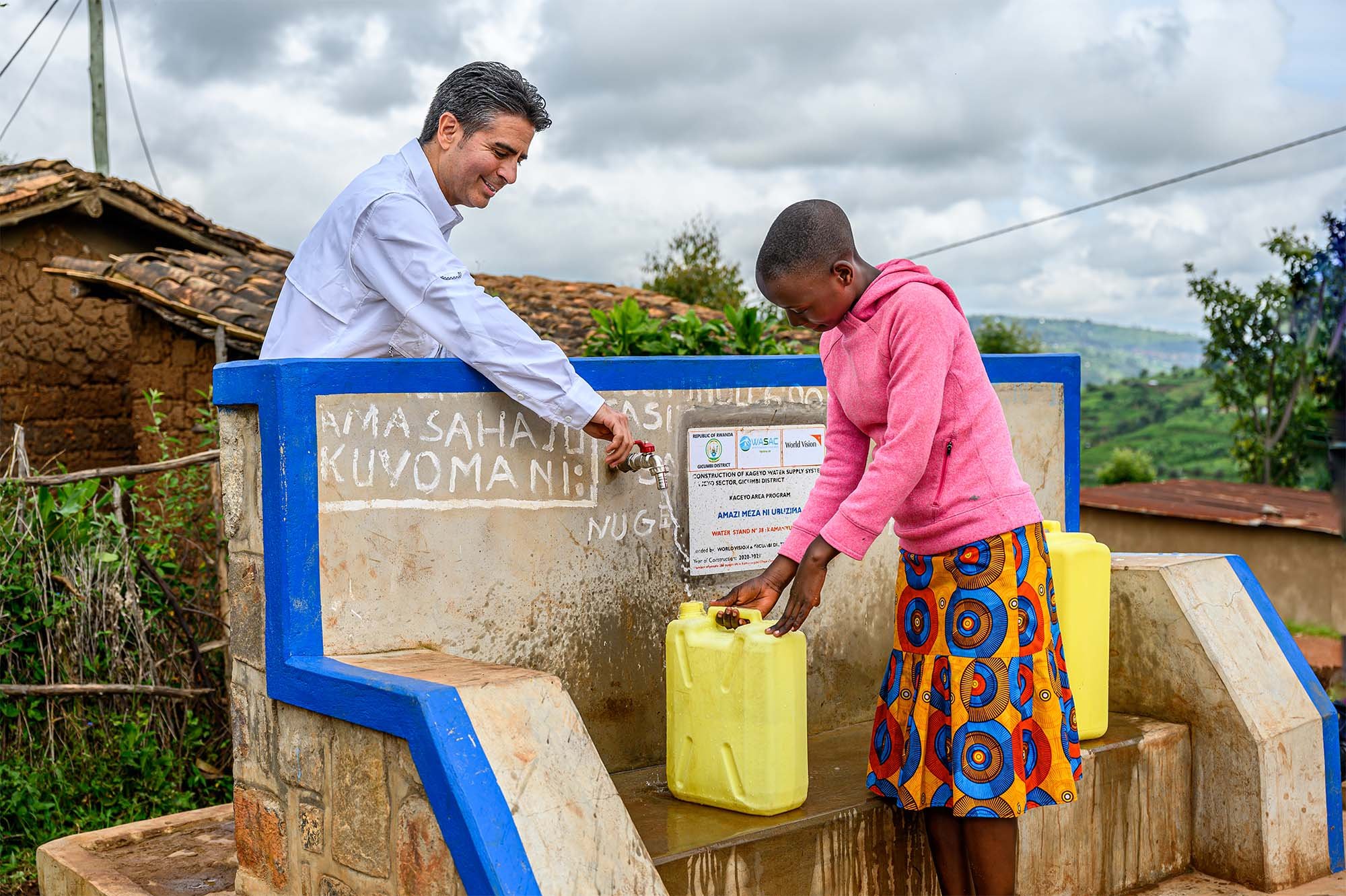 In Rwanda, a man smiles and stands behind a water tap, dispensing water into a container that a teenage girl is holding.