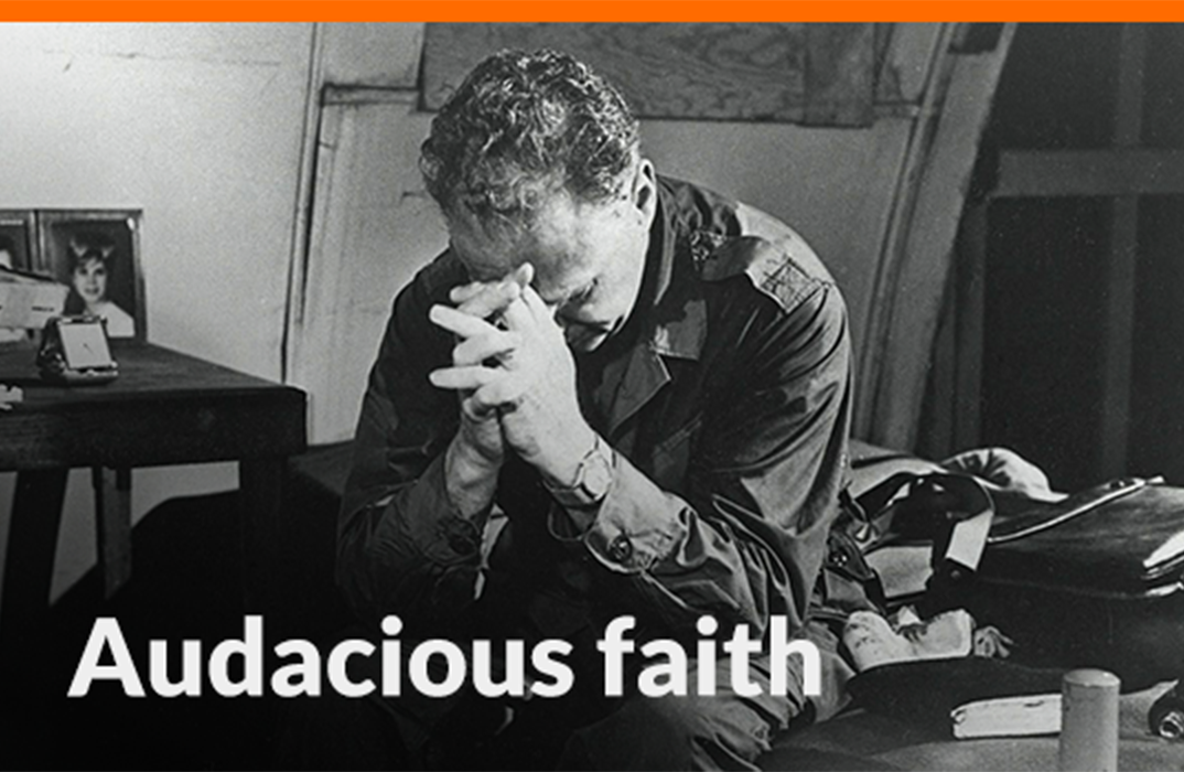 A black and white photo of a man with his head bowed, praying from the edge of a bed