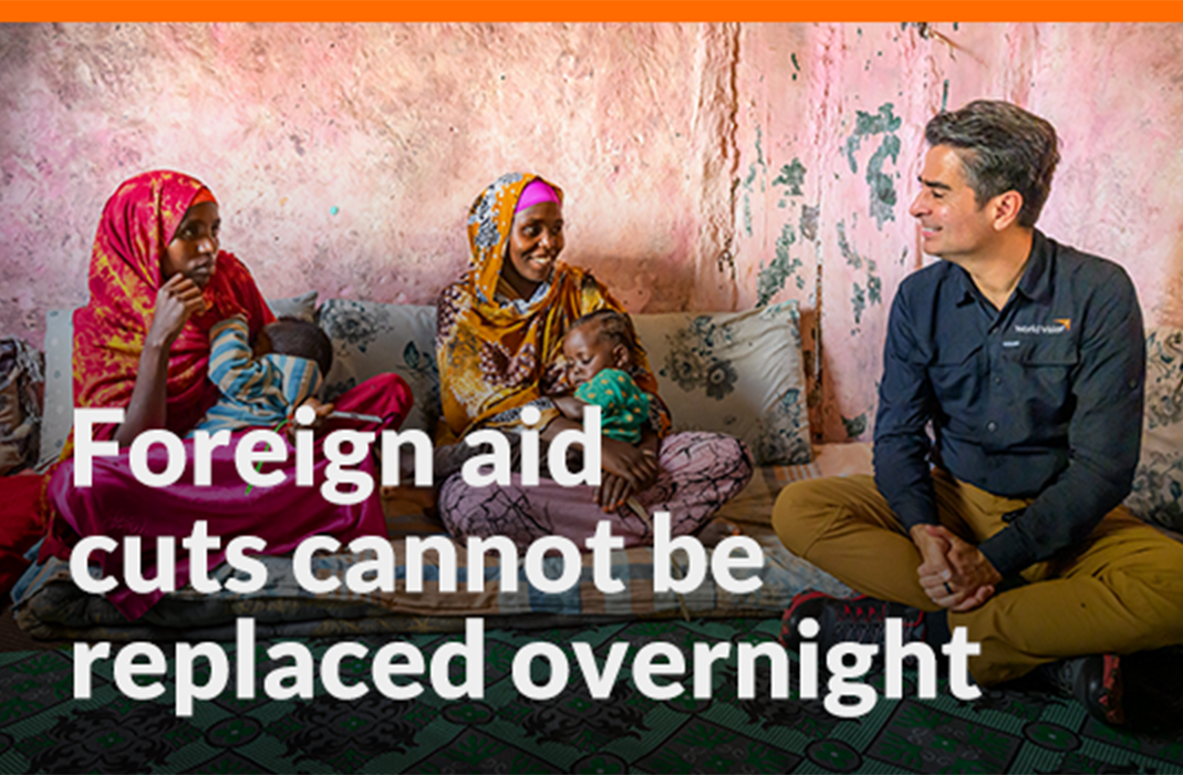 A man in a World Vision button-up shirt sits on the ground with two Ethiopian women, each holding a young child on their lap