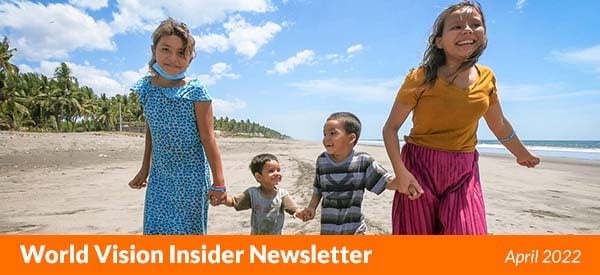 Four children from El Salvador run along a sandy beach with palm trees and blue sky behind them.