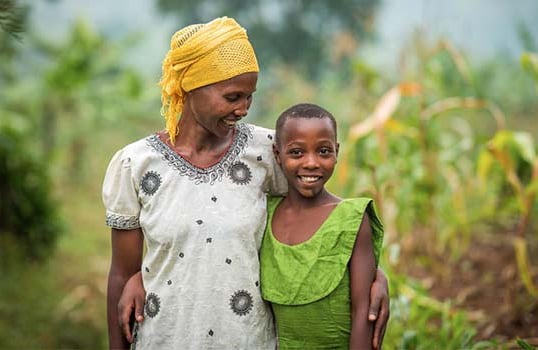 A Rwandan woman and younger girl stand together in a field. The older woman, in a white tunic and yellow headscarf, smiles down with pride with her arm around the girl, who’s wearing a bright green dress.
