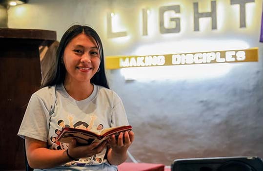 A teen girl from the Philippines smiles while holding open a Christian book at her church; an illuminated sign behind her shows the words “light” and “making disciples.”