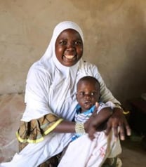 Zara, a mother in Niger, holds her son, Sani, on her lap, as she smiles. Zara is dressed in all white, and Sani wears a blue shirt and the remains of a nutritious meal on his face.
