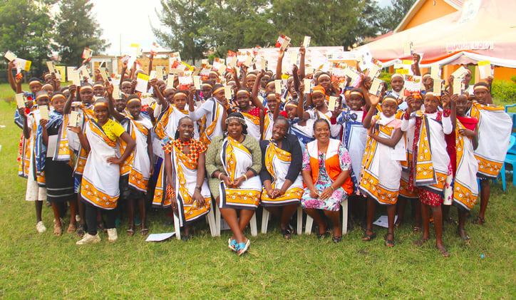 A large group of Kenyan girls and women sitting or standing outside hold certificates in the air and smile.