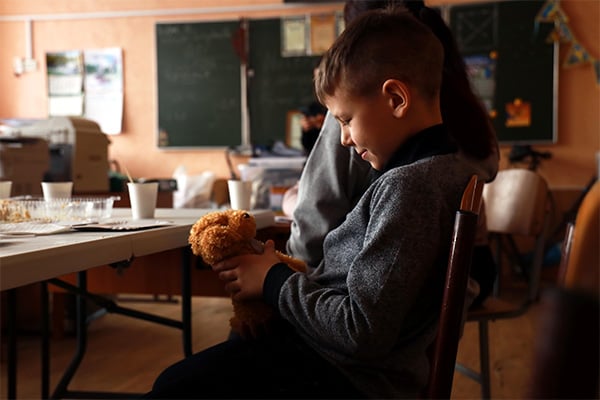 A young Ukrainian boy sits in a chair and smiles at a new teddy bear he’s just received. 