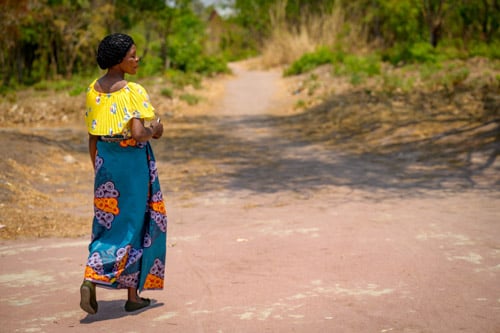 A woman wearing a yellow blouse walks down a dirt road in Zambia.
