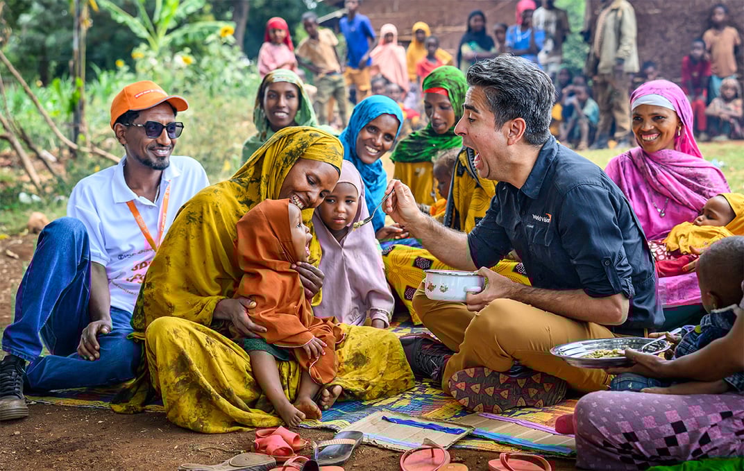 Edgar Sandoval sits on the ground next to a mother holding a young girl, feeding the child a bite of food with a spoon.
