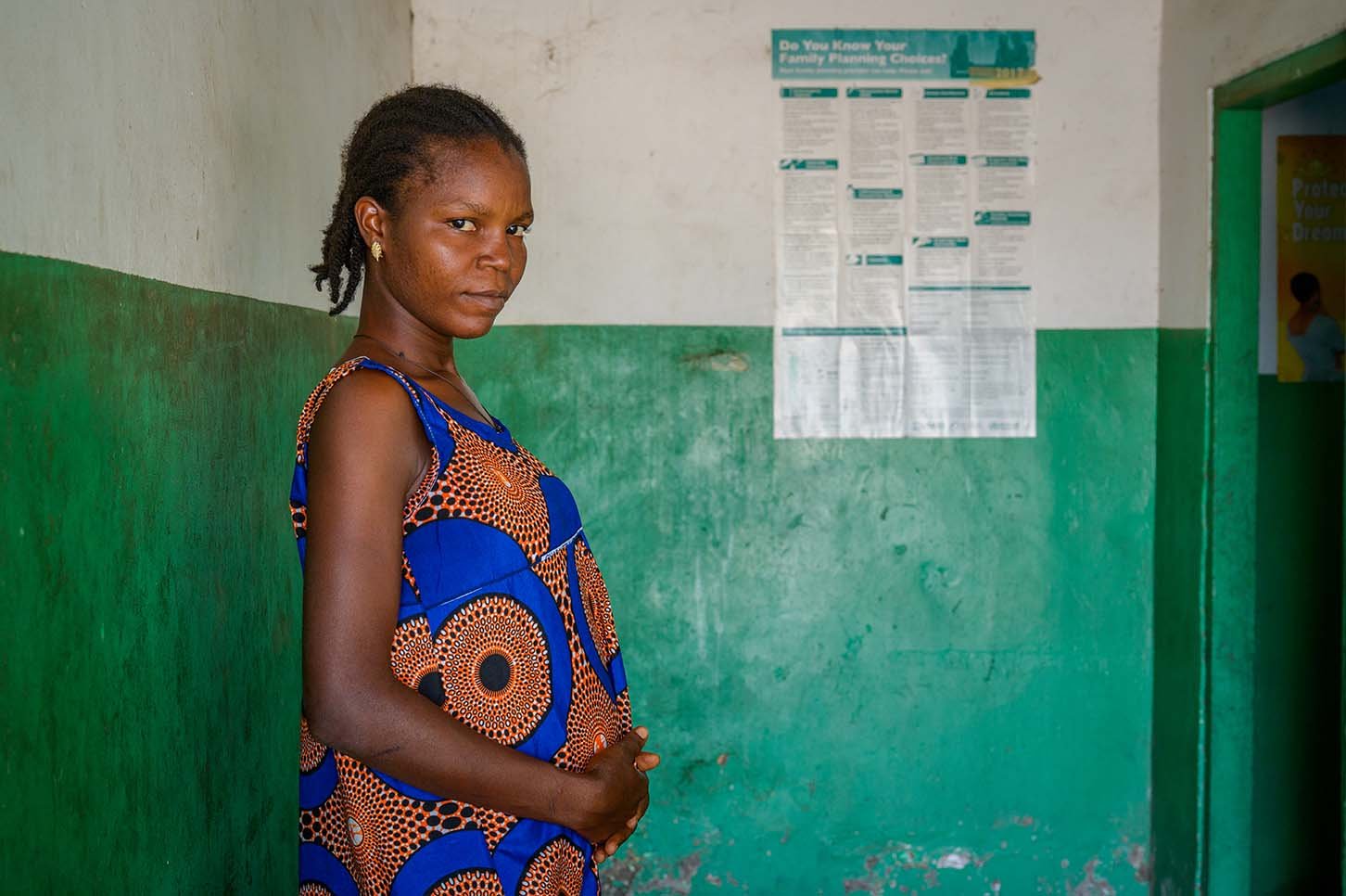 A pregnant woman in a dress holds her abdomen with a stoic expression.