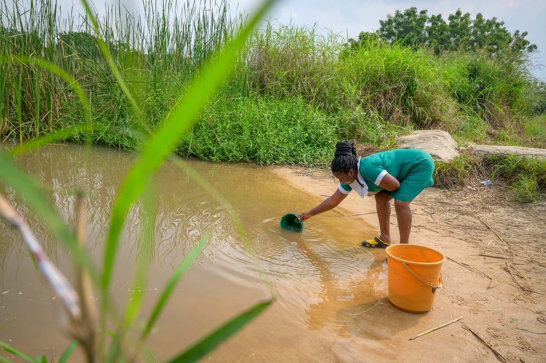 A woman scoops up dirty water from a sandy bank.