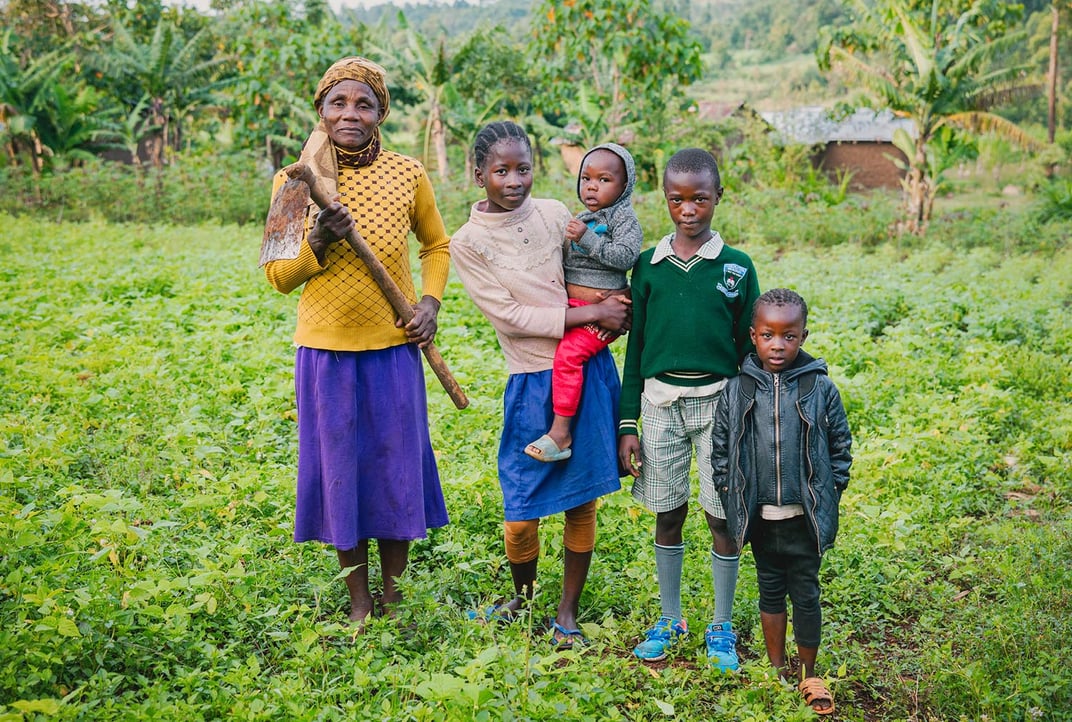 An older woman stands in a green field holding a tool; next to her are four children.