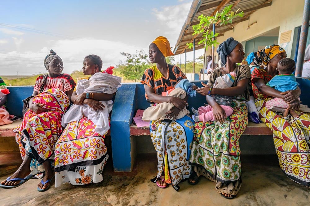 Five colorfully dressed women sit next to each other, each holding a baby.