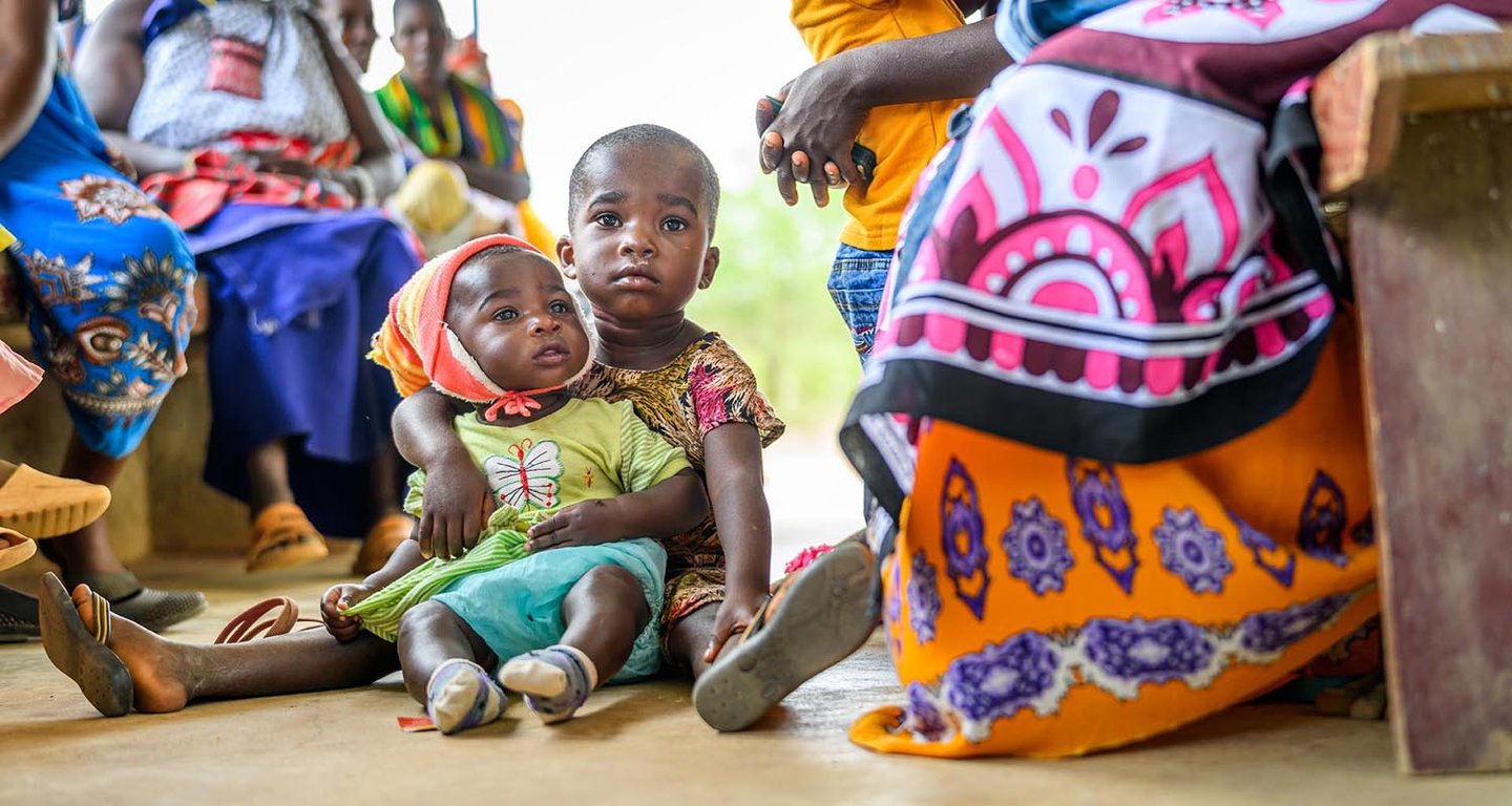 A child sits on the floor holding an infant, surrounded by seated women.