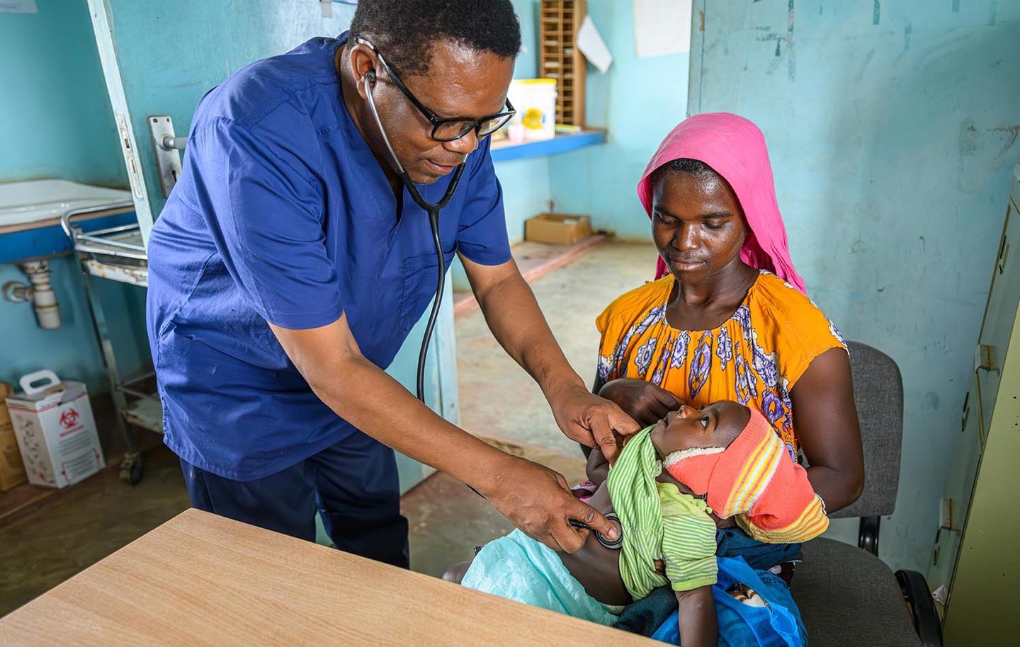 A man with a stethoscope attends to a young child being held by a woman in a clinic.