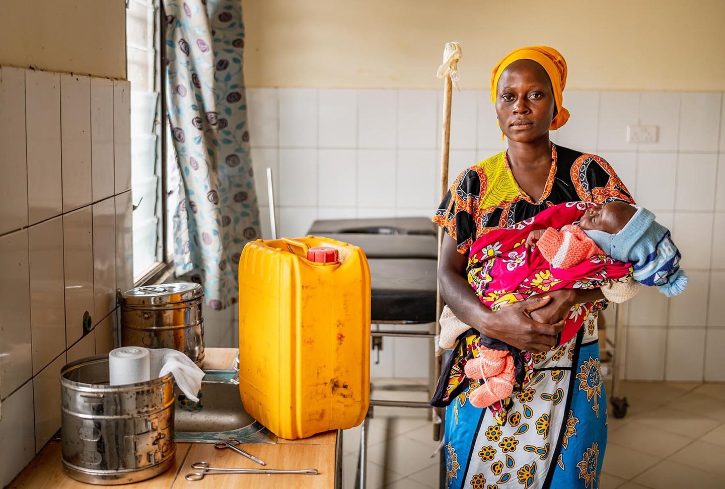 A woman holding a baby in a tiled room stands solemnly next to a large container used for carrying water.