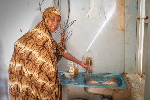 A Nigerien woman wearing a colorful patterned headscarf happily shows the clean water available in all the rooms of a health center. 