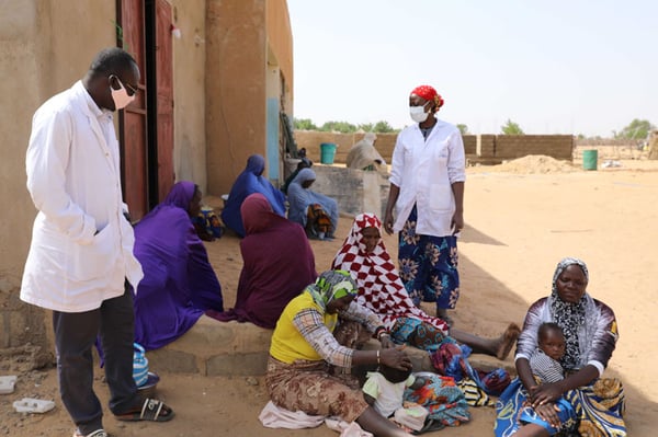 A doctor and a nurse in white coats talk to Nigerien mothers and children who are sitting in the dry and dusty sand outside a health center.
