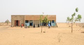 A health center rests in the midst of a sand dune, with a few sparse trees dotting the entrance and a few people walking toward it.
