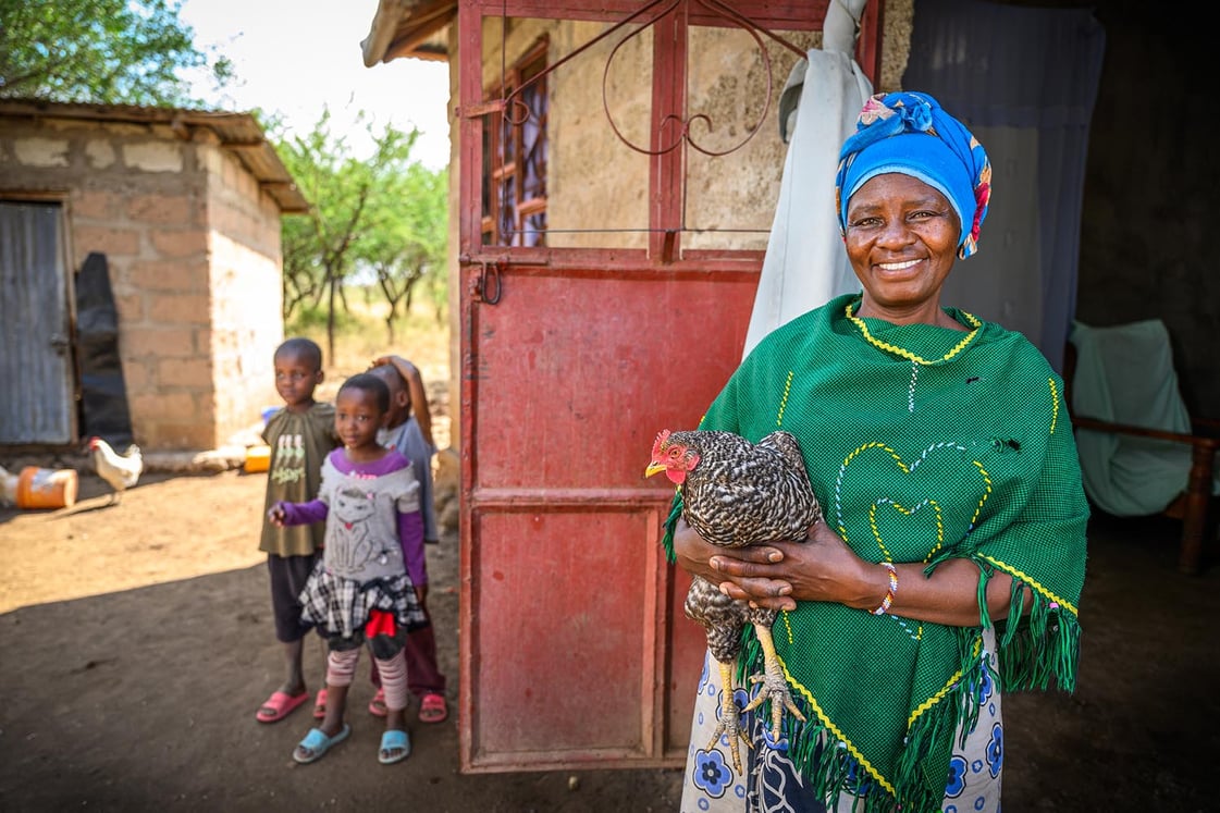A woman is smiling and holding a chicken standing outside of a house and next to a child.