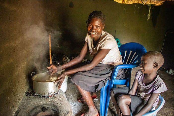 A mother in the Bidibidi settlement smiles as she prepares a meal from food aid next to her daughter.